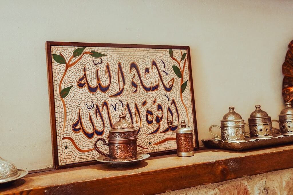 A paiting with Arabic letters on a wooden shelf with metal cups at one of the best Syrian tea rooms in London that we will take you to on the Syrian Heritage Tour.