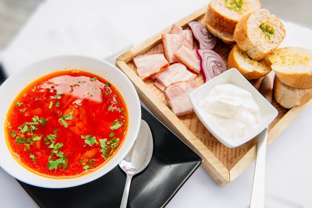 A bowl of traditional Ukrainian borsch next to a tray with bacon, onions, garlic bread and sour cream.