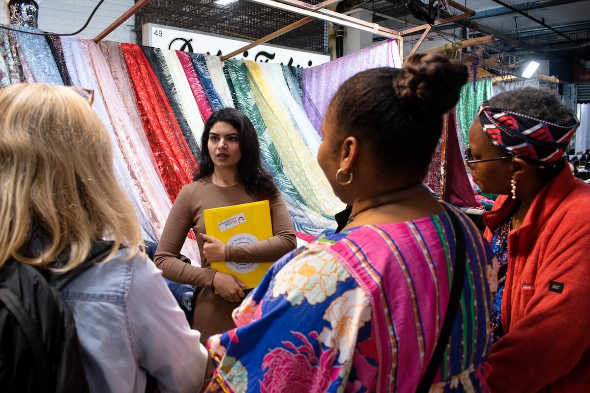 Fatima talking to guests in front of draped sheer and embroidered fabrics at a market textile stall in East London