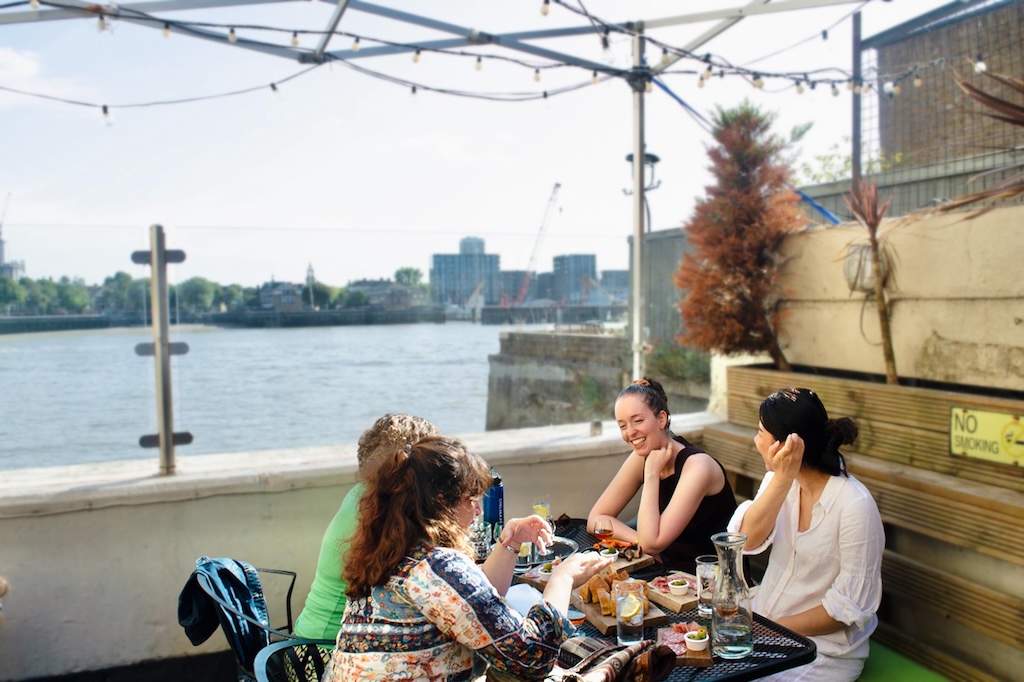 Florbela and her group of guests in the Iberian tour of London sitting at a riverside restaurant, trying Port wine and Portuguese tapas while sharing stories of the Iberian heritage in London.