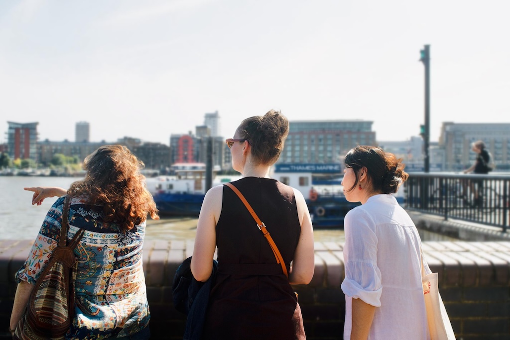 The Iberian tour guide, Florbela, and two female guests standing on the banks of the river Thames, with Florbela pointing at something outside of the frame of the image.