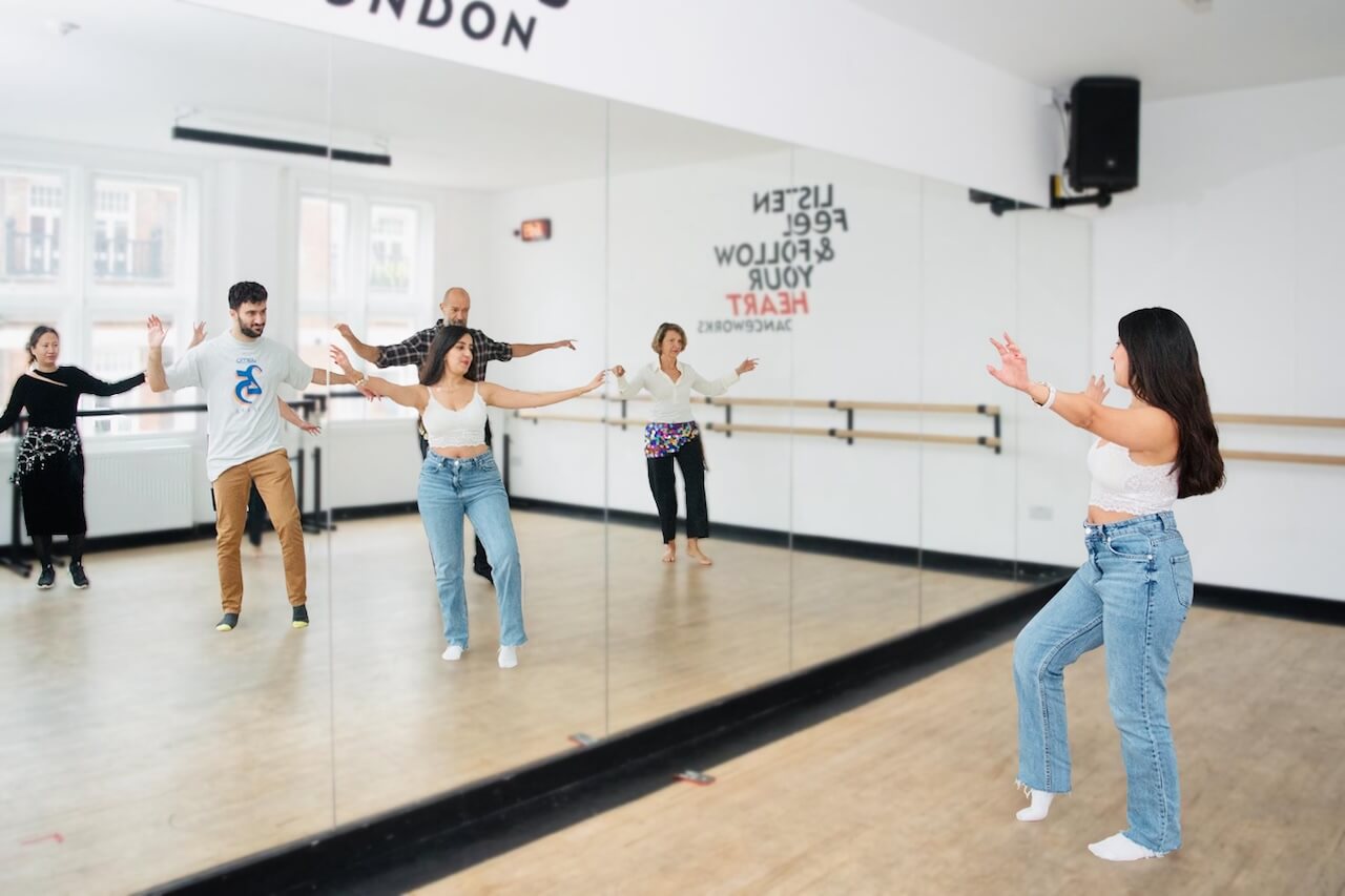 Amal leading a belly dancing class at Danceworks London, standing in front of a mirror wall while participants follow her movements with arms raised, practicing traditional dance steps together.