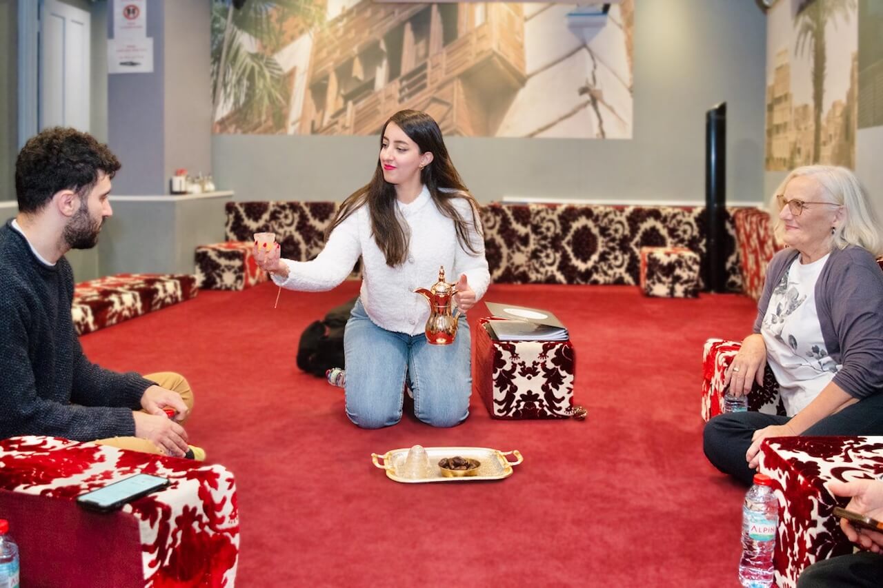 Amal kneeling on a red carpet, pouring traditional Arabic coffee for a guest as part of a Saudi hospitality experience in London. Two participants sit around her, smiling and engaging in the cultural ritual.