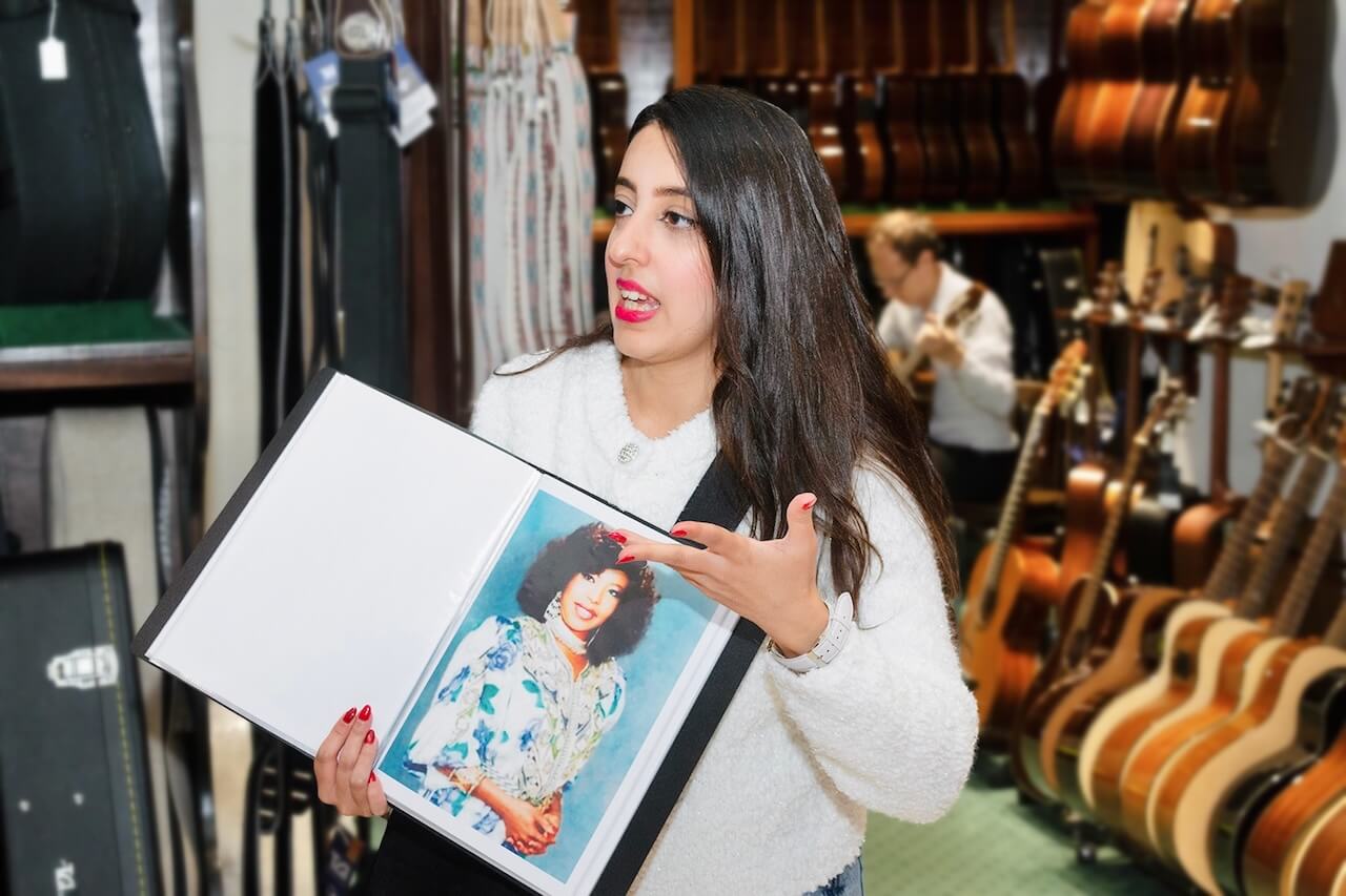Amal holding up a folder with a photo of a famous Saudi female musician, explaining Saudi culture inside a music shop surrounded by guitars.