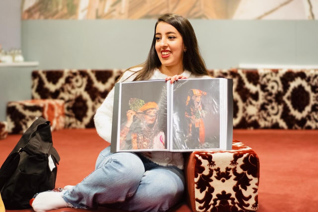 Amal sitting on the floor, holding open a folder with photographs of traditional Saudi clothing and smiling while sharing stories about cultural heritage.
